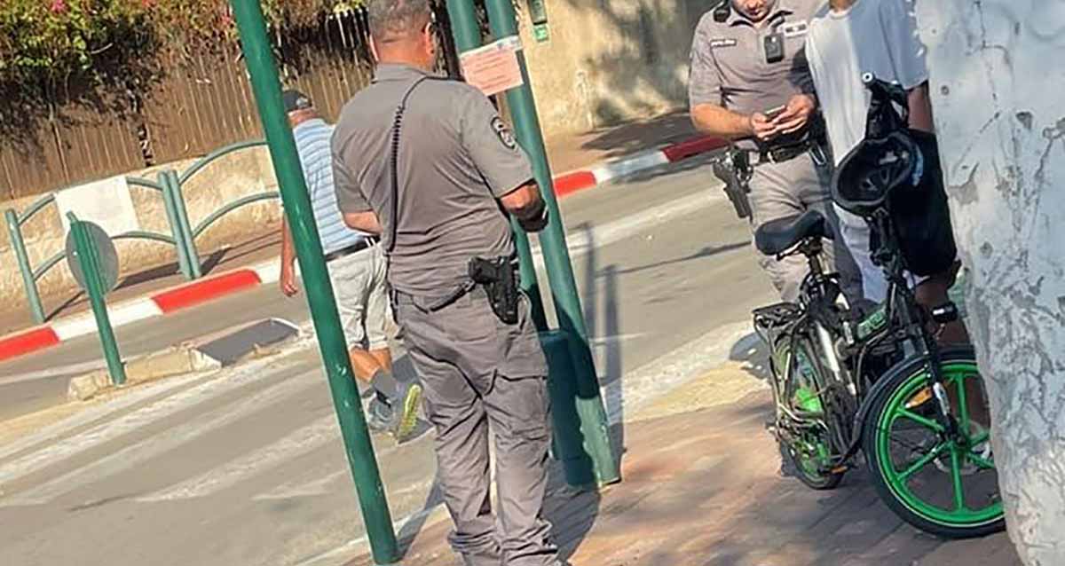 אכיפת קנסות קורקינטים ברמת השרון. שני שוטרים ליד קורקינט על מדרכה. בכביש מעבר חצייה והולך רגל חוצה את הכביש. משמאל גדר עץ של בית.