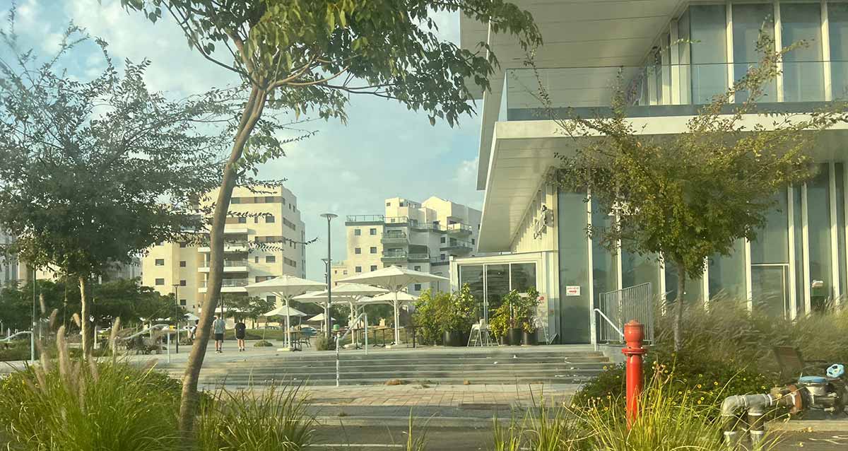 הקניון ובתים של שכונת נווה זמר ברעננה. בצד ימין נראה חלק מהצד הצר של הקניון , ובחזיתו שטח סגור בלוחות זכוכית ולפניו שמשיות עם שולחנות של בר. ברקע בתי מגורים של 7, 8 קומות. משמאל עצים ומתקני שעשועים לילדים.