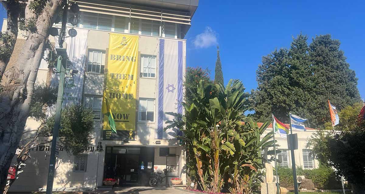 בניין עיריית רמת השרון. בניין בן 4 קומות. הכניסה בצד ימין. משמאל לכניסה על הקיר כתוב: עיריית רמת השרון. מהקומה הרביעית ועד לקומה הראשונה פרושה יריעה צהובה ועליה הכיתוב באנגלית: BRING THEM HOME NOW. מימין לשלט זה פרוש דגל המדינה באותו אורך. ומשמאל לו פרוש דגל רמת השרון. לפני הבניין עצים. מימין לבניין בחזית שלושה דגלים על תרנים. ובצד שמאל של הבניין נמצא מבנה חד קומתי ואחוריו עץ ענק.