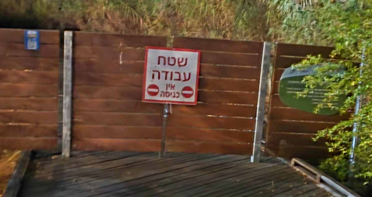 הכניסה לשביל ה"דק" חסומה על ידי גדר מלוחות עץ בצבע חום. על הנגדר שלט עם שני עיגולים המסמנים אין כניסה, וכיתוב: שטח עבודה אין כניסה.