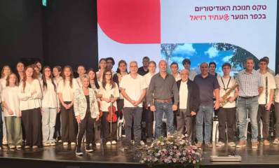 טקס חנוכת האודיטוריום בכפר הנוער דוד רזיאל. על הבמה עומדים תלמידים ומורים ולפניהם זר פרחים גדול. מעליהם שלט עם הכיתוב: טקס חנוכת האודיטוריום בכפר הנוער @עתיד רזיאל.