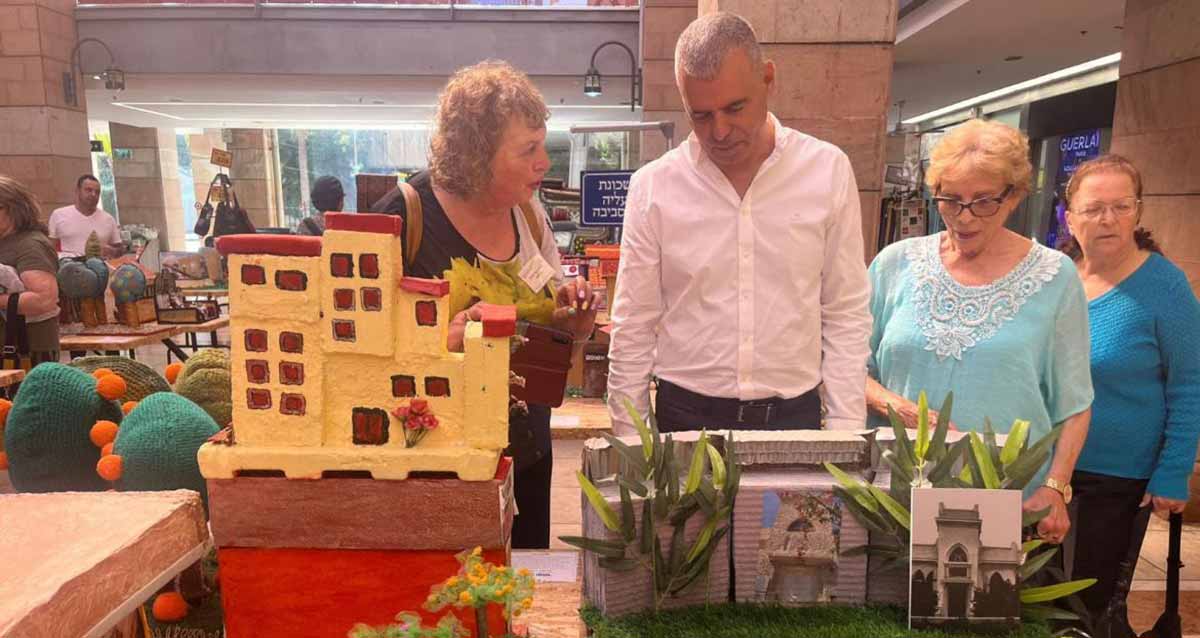 תערוכה אמנותית של ותיקי העיר כפר סבא. ראש העיר ותושבים ותיקים מתבוננים בעבודות האמנות, בתוך אולם בו העבודות מונחות על שולחנות.