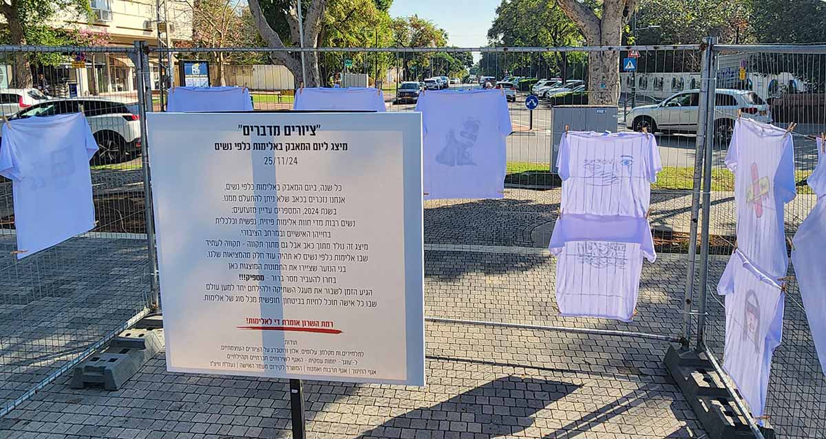 מיצג ציורים מדברים ליום המאבק באלימות כלפי נשים בתאריך 25/11/24. במרכז התמונה שלט עם הכותרת "ציורים מדברים", והסבר על המאבק והמיצג. מסביב לשלט תלויות על גדר חולצות טישרט לבנות ועליהן ציורים.