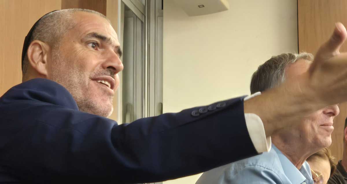 יעקב קורצקי מניף ידו הימנית קדימה. צילום פלג גוף עליון. לבוש ז'קט כחול וחולצה לבנה.