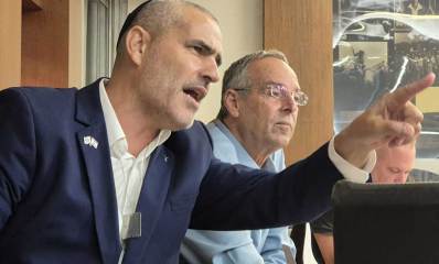 יעקב קורצקי מניף ידו השמאלית קדימה ומצביע עם אצבע קדימה. צילום פלג גוף עליון. לבוש ז'קט כחול וחולצה לבנה. לידו ראש העיר איציק רוכברגר ממושקף בחולצה בצבע תכלת.