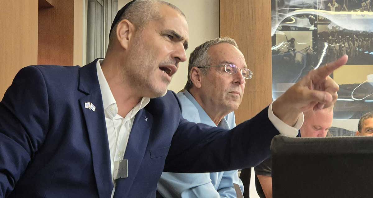 יעקב קורצקי מניף ידו השמאלית קדימה ומצביע עם אצבע קדימה. צילום פלג גוף עליון. לבוש ז'קט כחול וחולצה לבנה. לידו ראש העיר איציק רוכברגר ממושקף בחולצה בצבע תכלת.