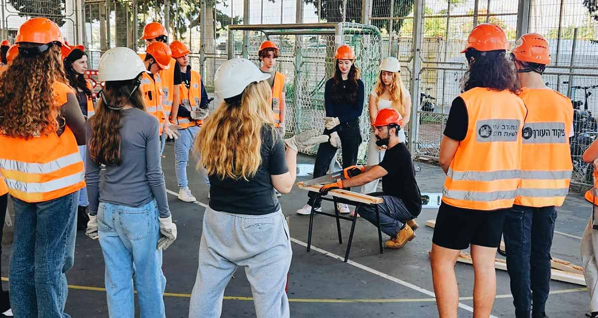 מחלצים צעירים בתיכון חדש. תלמידות ותלמידים עם ווסטים וקסדות של פיקוד העורף עומדים סביב מדריך האוחז בידו משור וחותך פרופיל.