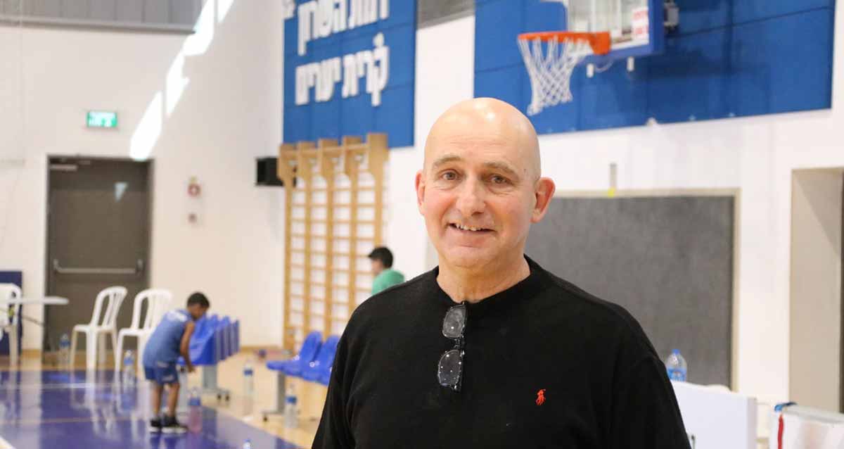 אריק דורון מנכ"ל אגודת ספורט רמת השרון. בחולצה שחורה באולם ספורט. מאחוריו סל של כדורסל. על הקיר מאחוריו כיתוב: רמת השרון קרית יערים וסולם התעמלות על הקיר מתחת לכיתוב. שני ילדים מאחוריו.