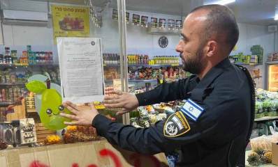 צו סגירה לחנות ירקות ברמת השרון. שוטר מדביק את הצו על חלון הזכוכית של החנות. ברקע פנים החנות עם ירקות, פירות ושימורים.
