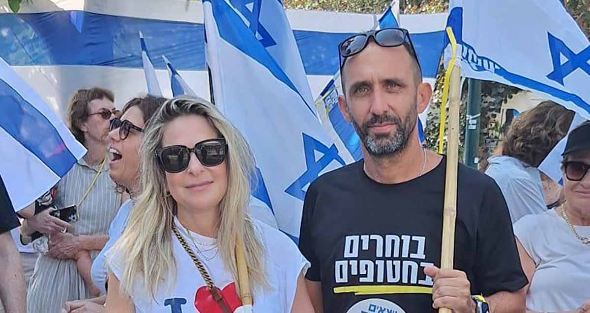 גיא קלנר וסיון שמרי דהן בהפגנה עם דגלי המדינה. גיא בחולצה שחורה עם כיתוב: בוחרים בחטופים. סחון עם משקפי שמש כהים, חולצה לבנה ועליה האות I באנגלית ולב אדום. מאחוריהם מפגינים.