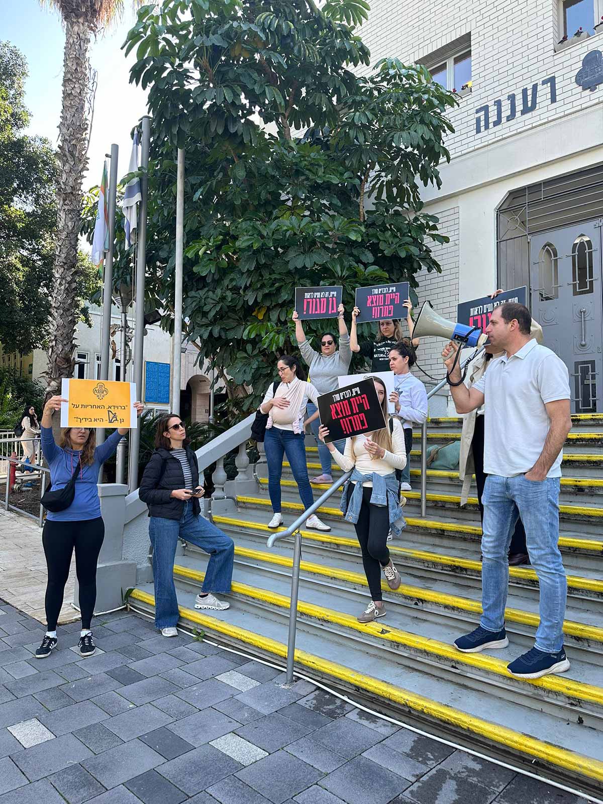 הפגנת אמהות לילדי גן נבל משכונת נווה זמר מחוץ לבניין עיריית רעננה. אמהות עם שלטים על מדרגות הכניסה לבניין העירייה. וגבר אחד עם מיקרופון נייד.