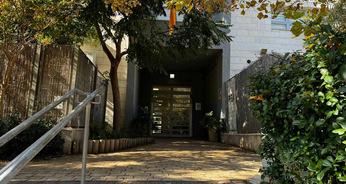 גן נבל בשכונת נווה זמר ברעננה. צילום של הכניסה הגדולה לבניין. עץ משמאל לכניסה.מימין ומשמאל גדרות של במבוק מסביב לחצרות.