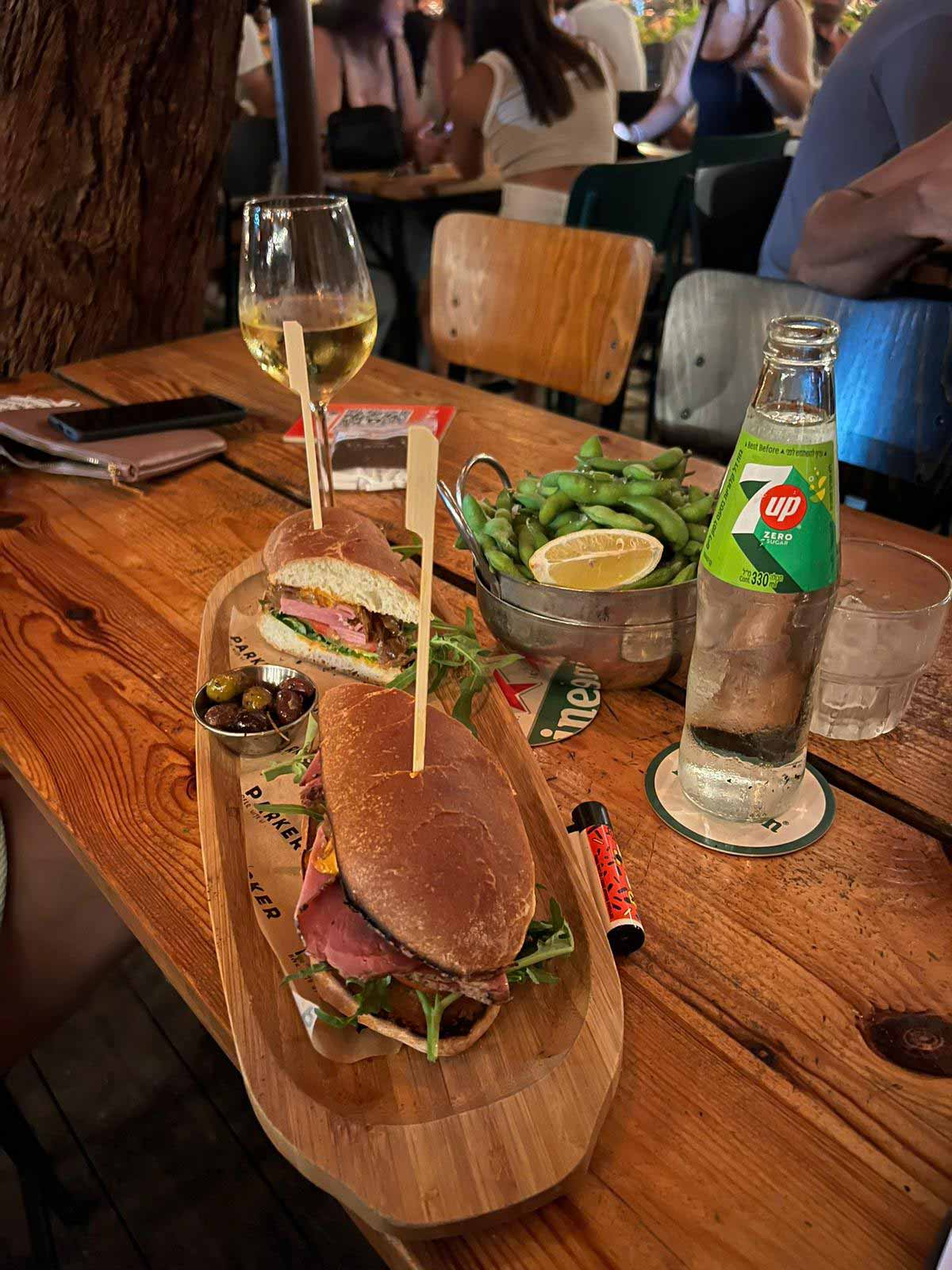 פארקר בר מסעדה בפארק הרצליה. תמונה של שולחן ועליו בצלחת אליפטית מעץ, מונח כריך חצוי לשניים ולידו צלוחית זיתים. ליד הצלחת צלוחית עם סלט, בקבוק שתייה "סבן אפ" וכוסית עם יין לבן.