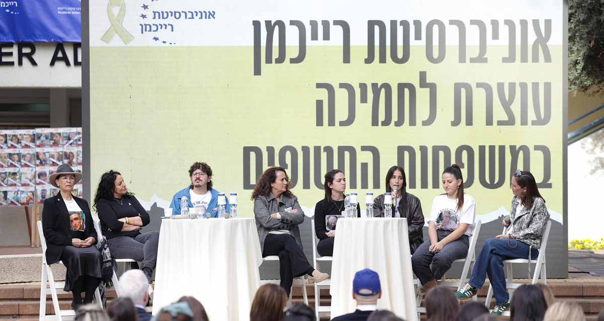 עצרת תמיכה במשפחות החטופים באוניברסיטת רייכמן. על במה יושבים סטודנטים ומשפחות חטופים ולפניהם שני שולחנות עגולים עם מפות לבנות. מאחוריהם פלקט ענק עם סמל האוניברסיטה ושמה של העצרת.