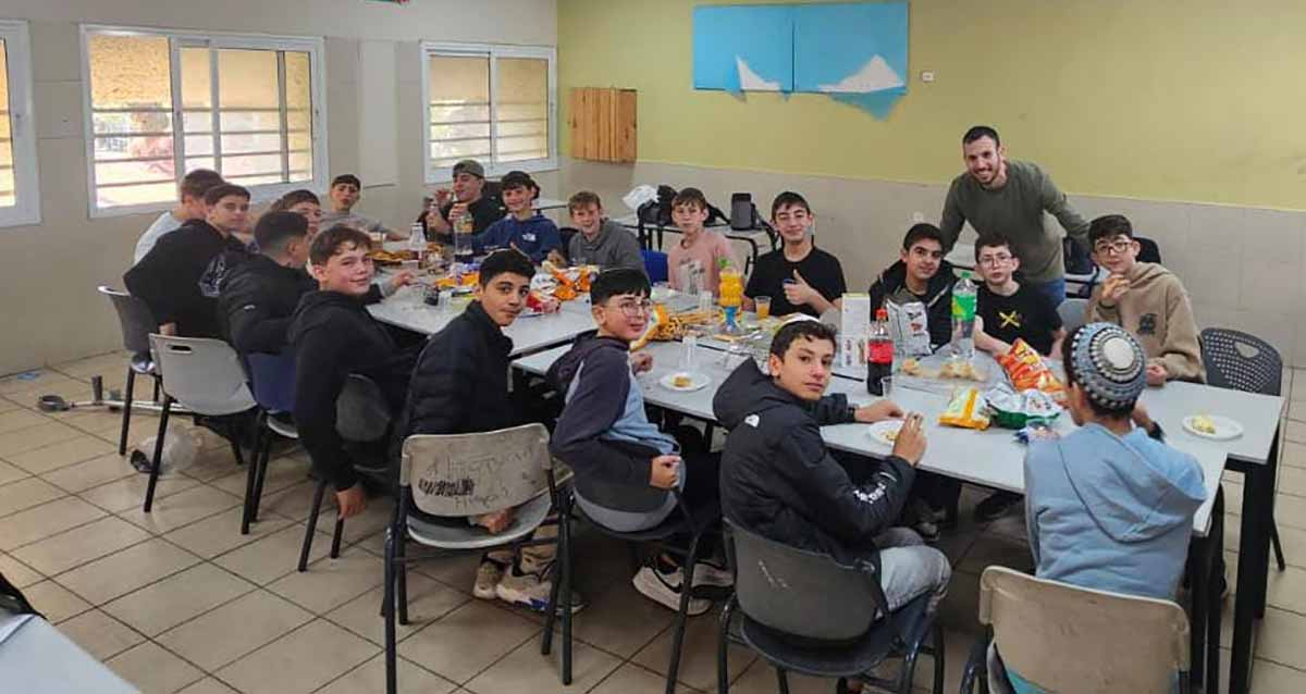 התלמידים ערכו סעודת הודיה למורה שחזר ממילואים באמית כפר בתיה. סביב שולחן עם עוגה, חטיפים ושתייה יושבים התלמידים והמורה עומד מאחוריהם.
