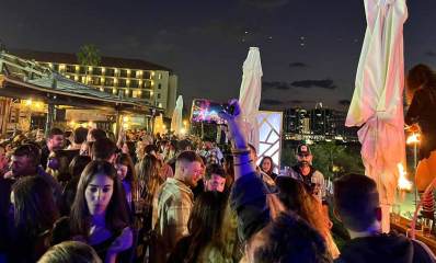 ים בר בהרצליה. ברחבה לפני הבר בשעת חשיכה, המוני צעירות וצעירים עם כוסות. ברקע הבר ומלון.