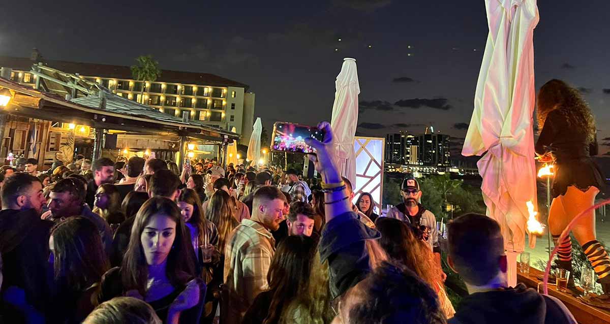 ים בר בהרצליה. ברחבה לפני הבר בשעת חשיכה, המוני צעירות וצעירים עם כוסות. ברקע הבר ומלון.