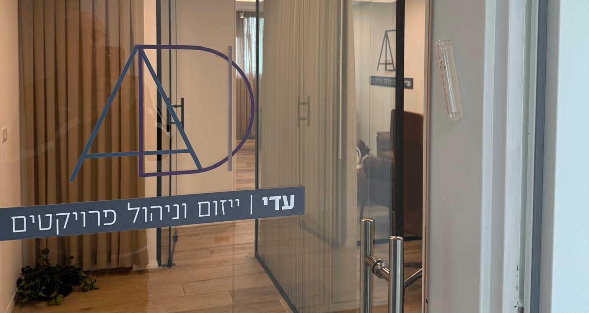 דלת זכוכית משרדית ושקופה ופנים משרד עם רצפת פרקט. על הדלת יש לוגו עם האותיות AD ועל הדלת כתוב: עדי | ייזום וניהול פרוייקטים