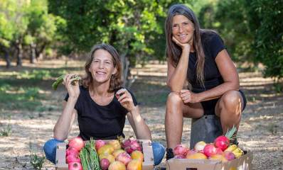 ענת רשף גלעד וענת קרץ עם קרטונים מלאים בפירות, אחת יושבת על הרצפה והשנייה על דלי, בשטח חשוף ומאחוריהן עצים.