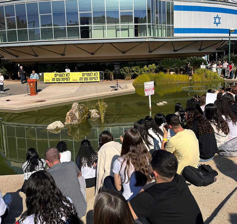 רפי סער נואם בעצרת למען שחרור החטופים. בקהל תלמידי שכבת י. לפניו בריכת נוי, מאחוריו שלט בו כתוב: לא לומדים לחיות עם זה. על הבניין דגל ענק של ישראל