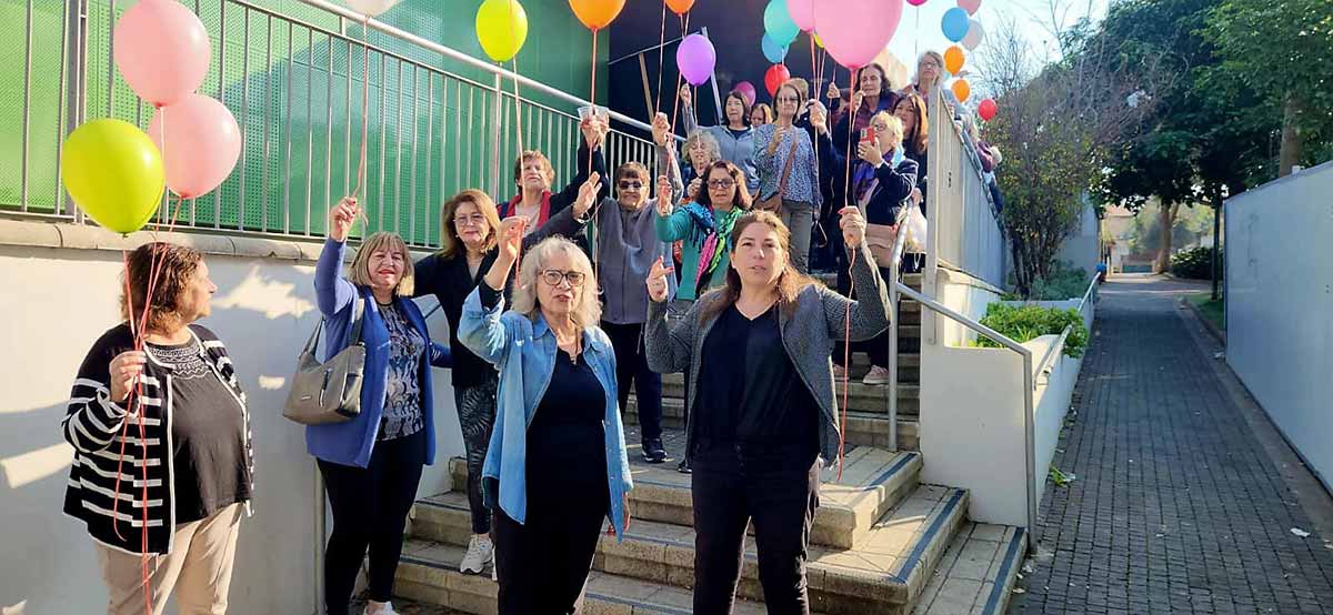 קבלת פנים לסבתא של לירי אלבג. חבורה גדולה של נשים עומדות עם בלונים צבעוניים בפתח של בניין על גרם המדרגות.