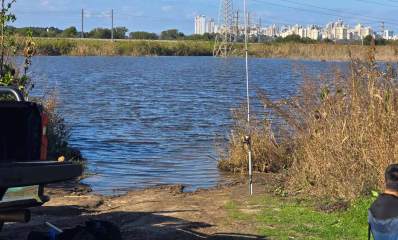 פארק יקום. תמונה של אגם מים כחולים, עשבים וצמחים בשתי הגדות. בגדה הקרובה בצד ימין יושב אדם על כסא מבד, ומכוניתו בצד השני. בהמשך מסילת ברזל ושורה של בתים באופק.
