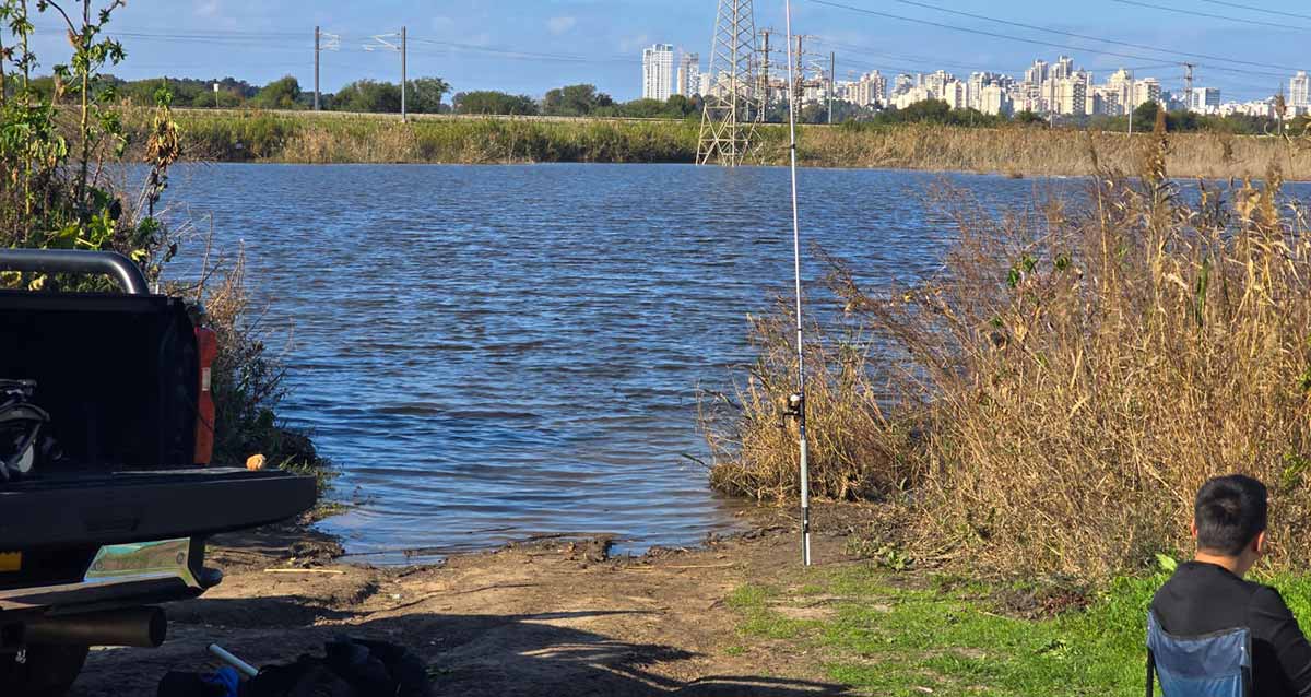 פארק יקום. תמונה של אגם מים כחולים, עשבים וצמחים בשתי הגדות. בגדה הקרובה בצד ימין יושב אדם על כסא מבד, ומכוניתו בצד השני. בהמשך מסילת ברזל ושורה של בתים באופק.