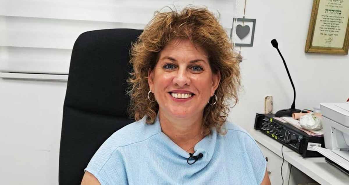 טלי תמרי גבע נהלת אגף החינוך היסודי בהרצליה. צילום פנים יושבת במשרד ולידה מדפסת על שולחן. על הקיר מאחוריה תמונה של לב ותעודת הוקרה.