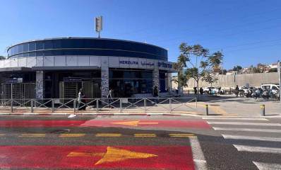 תחנת הרכבת הרצליה. בניין עגול של הכניסה לתחנת הרכבת. בקומת הקרקע כניסות ויציאות. הקומה השנייה חלונות זכוכית. על הגג אנטנה. מימין מכוניות בכביש, ואופניים ואופנועים על המדרכה.