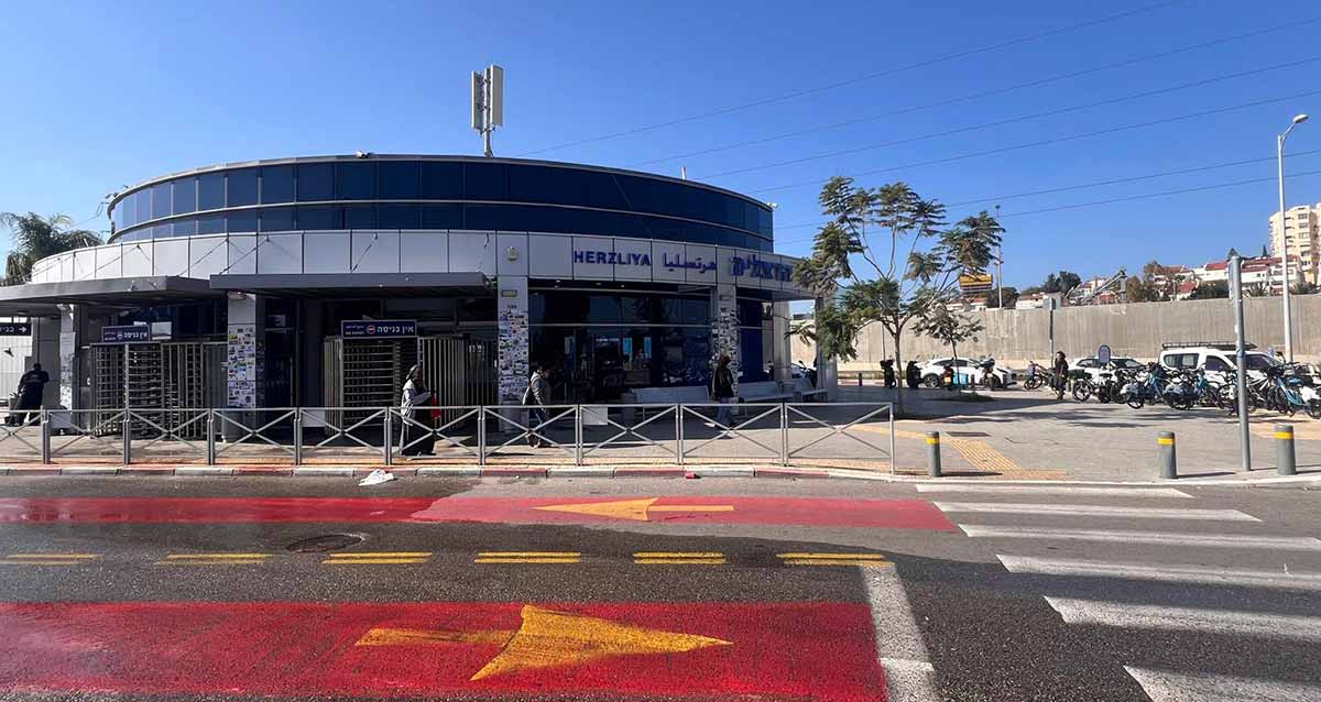 תחנת הרכבת הרצליה. בניין עגול של הכניסה לתחנת הרכבת. בקומת הקרקע כניסות ויציאות. הקומה השנייה חלונות זכוכית. על הגג אנטנה. מימין מכוניות בכביש, ואופניים ואופנועים על המדרכה.