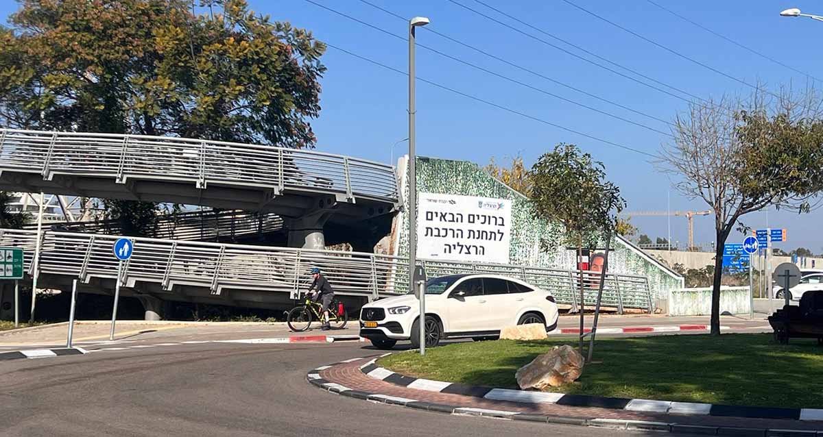 תחנת הרכבת הרצליה. בצד ימין כיכר ומכונית לבנה. ברקע גשרים להולכי רגל, ועל העלייה לגשרים שלט גדול עם כיתוב: ברוכים הבאים לתחנת הרכבת הרצליה.
