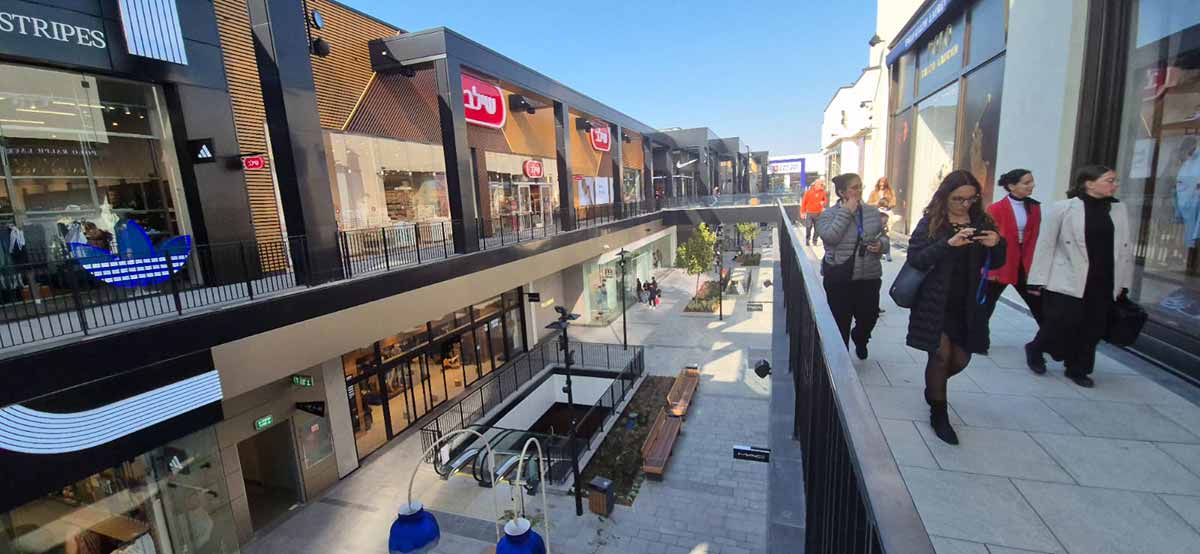 ביג פאשן גלילות. צילום מקומה שנייה ושתי הקומות בתמונה. חנויות בצד ימין ונשים צועדות לפני החנויות. ובצד שמאל שורת חנויות ובין שתי שורות החנויות נראית קומה ראשונה שבמרכזה מדרגת נעות כלפי מטה, ספסלי ישיבה וצמחייה.