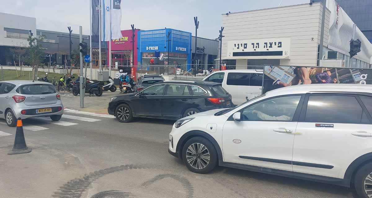 הכביש הפנימי שמוביל לביג פאשן גלילות. מכוניות פרטיות בכביש. ברקע מימין מבנה לבן עם הכיתוב: פיצה הר סיני. במרכז מבנה צבוע בכחול עם כיתוב באנגלית: B-FRESH. לידו מבנה צבוע באדום עם הכיתוב: אהרוניס. אופנועים חונים במדרכה לפני החנויות.