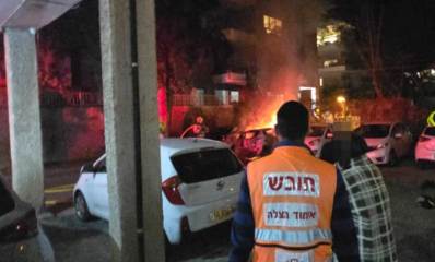 חובש איחוד הצלה עם הגב למצלמה צופה יחד עם כבאים ושוטרים ברכב עולה באש במגרש חנייה של בניין מגורים
