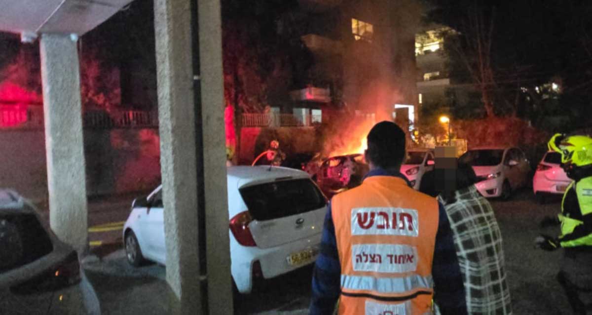 חובש איחוד הצלה עם הגב למצלמה צופה יחד עם כבאים ושוטרים ברכב עולה באש במגרש חנייה של בניין מגורים