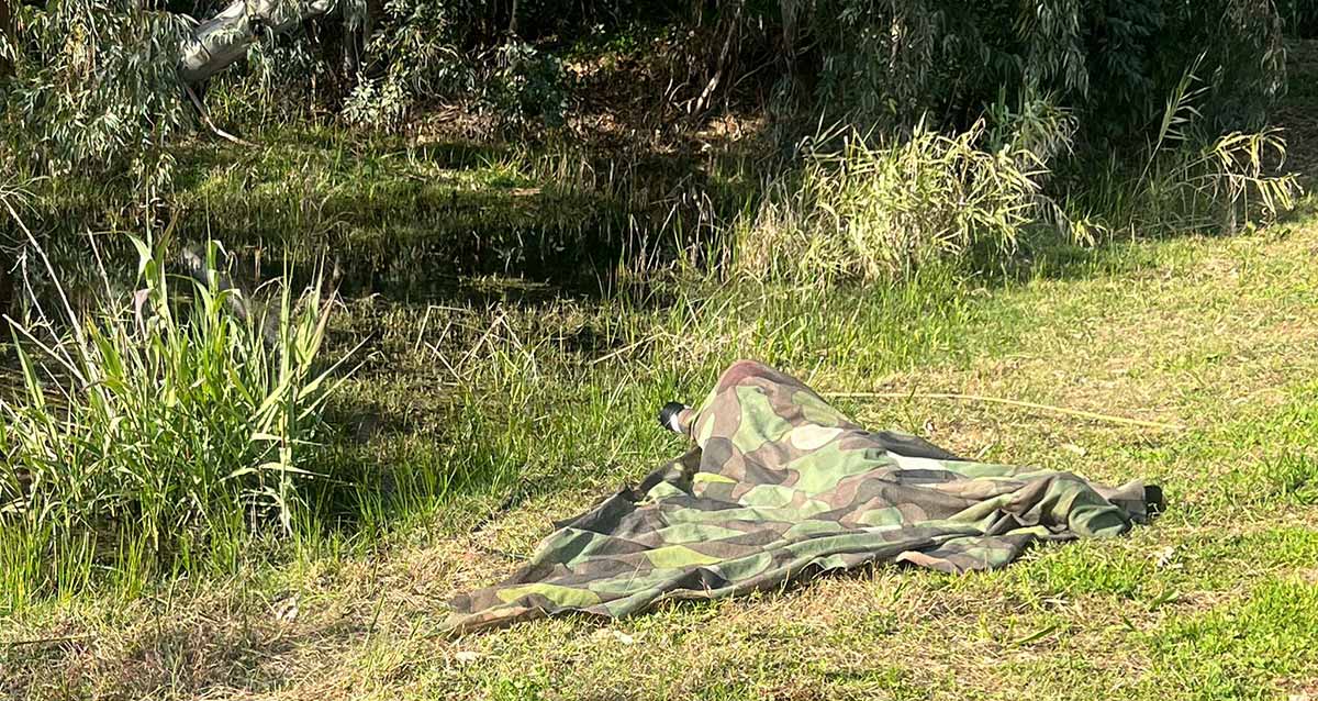 צלם ציפורים מוסווה מצלם עם עדשה גדולה עופות מים בפארק. הצלם שוכב על הדשא מכוסה ביריעת הסוואה.