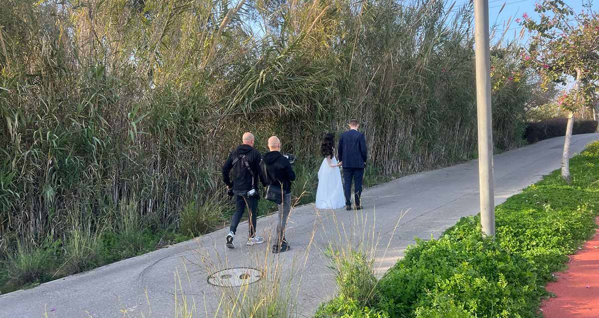 חתן וכלה מצטלמים בפארק הרצליה לקראת החתונה. החתן והכלה צועדים בשביל ואחריהם הצלמים. משמאל קני סוף, ומימין ירק ומסלול ריצה אדום.