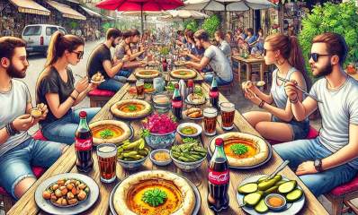 מסעדת חומוס. על המדרכה שולחנות ארוכים עם בחורות ובחורים יושבים ואוכלים. השולחנות עמוסים בקערות חומוס בצלחות עם מלפפונים וירקות, צלוחיות עם גרגירים, ולחמניות. בקבוקי קולה וכוסות בירה. הכביש משמאל חונות שתי מכוניות.