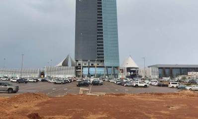 מגרש החנייה צמוד דופן אל ביג פאשן. מגרש עם הרבה מכוניות לכל רוחב התמונה. מאחורי המגרש נראה המגדל של ביג פאשן,. מימין מבנה חד קומתי ומשמאל גדר בטון. לפני המגרש שטח חולי.