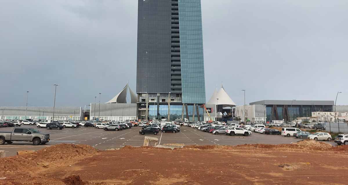 מגרש החנייה צמוד דופן אל ביג פאשן. מגרש עם הרבה מכוניות לכל רוחב התמונה. מאחורי המגרש נראה המגדל של ביג פאשן,. מימין מבנה חד קומתי ומשמאל גדר בטון. לפני המגרש שטח חולי.