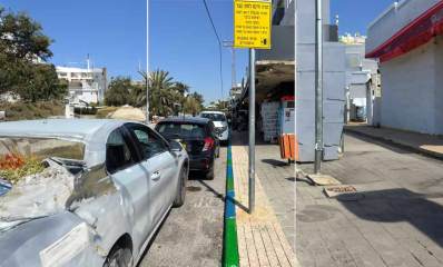 חנייה חינם לזמן קצר. שפת המדרכה בצבעים ירוק וכחול. במדרכה עמוד עם התמרור החדש של חניה חינם לזמן קצר. כלי רכב חונים משמאל, ומימין שורת חנויות..