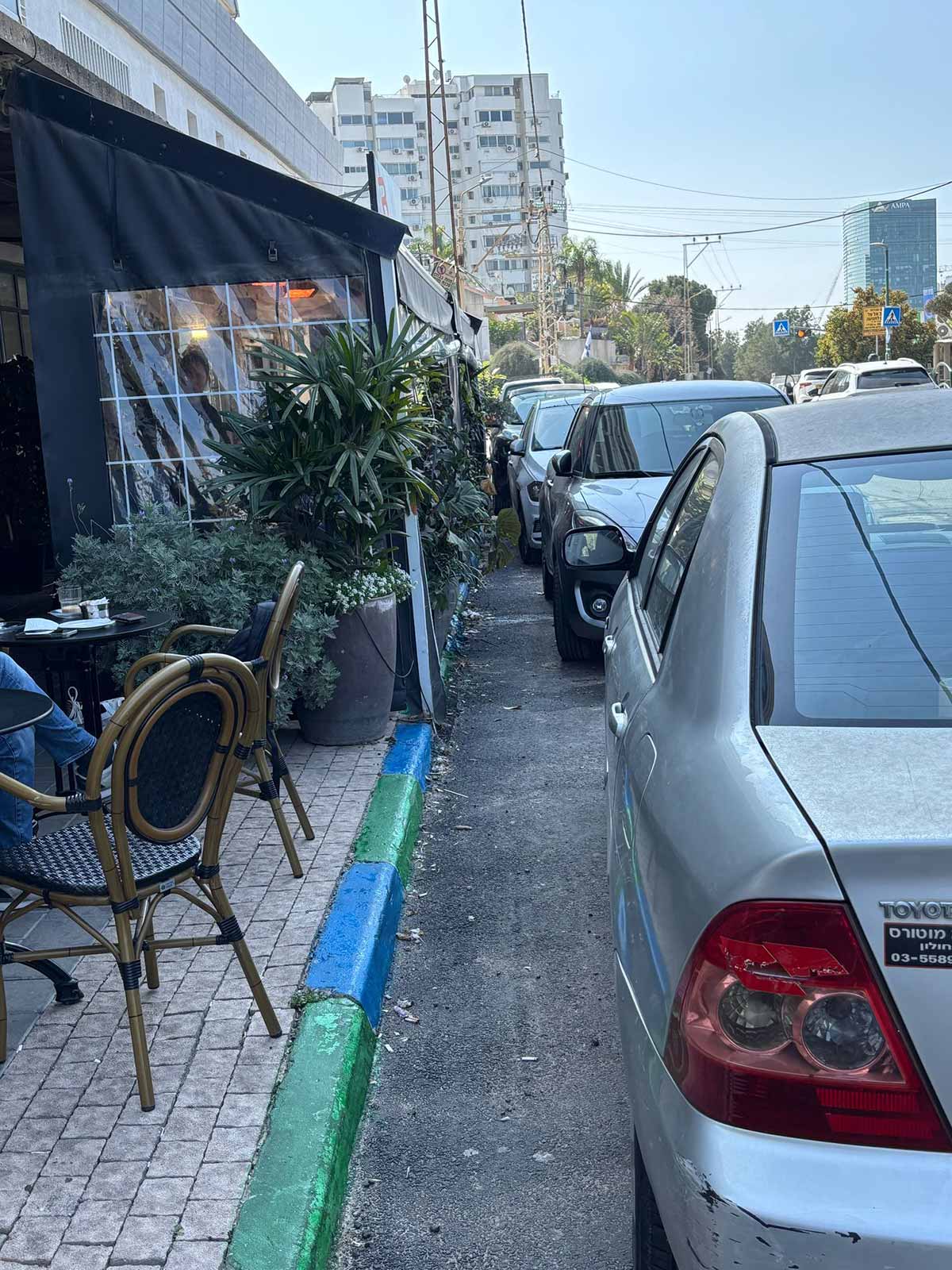 חנייה חינם לזמן קצר. שפת המדרכה בצבעים ירוק וכחול. כלי רכב חונים מימין, ומשמאל שולחן וכסאות של בית קפה.