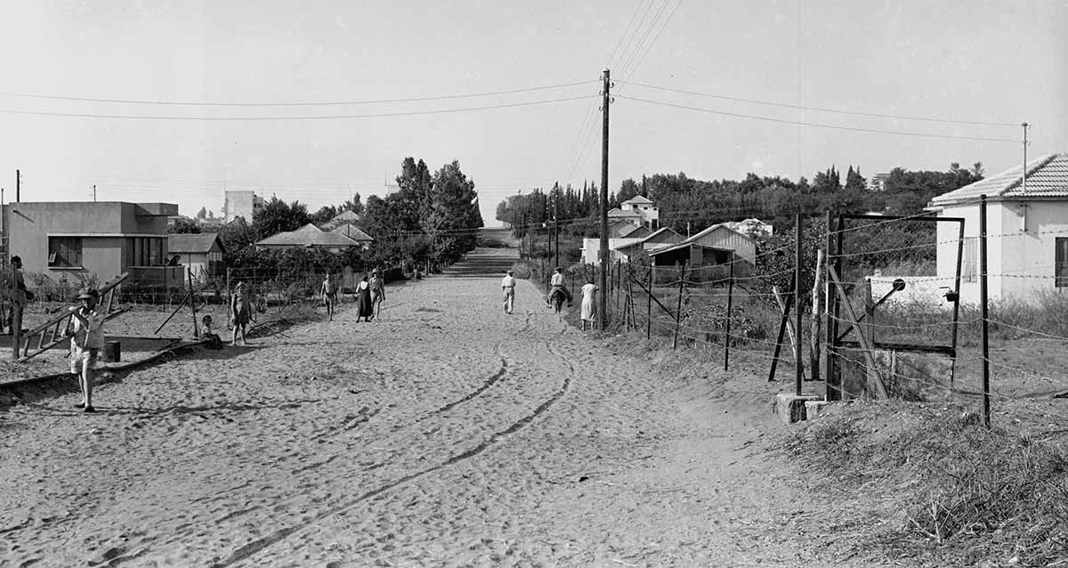 שכונת פועלים א', רחוב אהרונוביץ לפני סלילתו. חול ברחוב ואנשים ברחוב. אחד רוכב על חמור. מימין ומשמאל בתים חד קומתיים, חלקם עם גגות רעפים.