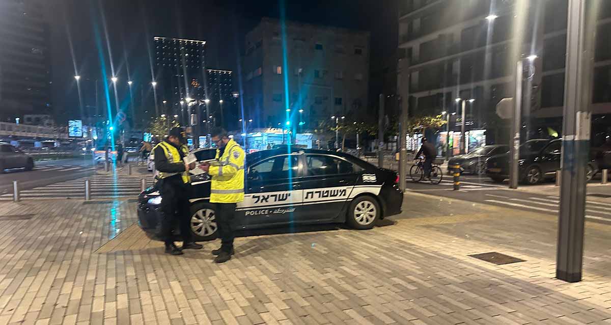 ניידת משטרה בפעילות. ניידת של משטרת ישראל חונה על מדרכה ולידה עומדים שני שוטרים במדים ובגד עליון צהוב. ברקע כבישים, מכוניות, חנויות ובניינים.