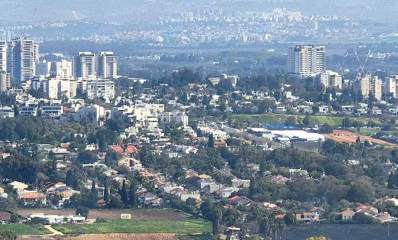 מבט על רמת השרון מכיוון מערב. השכונה הקרובה אלינו כולה בתים פרטיים, עם עצים רבים, ורובם עם גגות רעפים. במרכז התמונה מימין ומשמאל בתים רבי קומות ומגדלים. ברקע שכונות נוספות בתמונה מעורפלת.