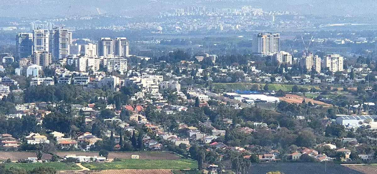 מבט על רמת השרון מכיוון מערב. השכונה הקרובה אלינו כולה בתים פרטיים, עם עצים רבים, ורובם עם גגות רעפים. במרכז התמונה מימין ומשמאל בתים רבי קומות ומגדלים. ברקע שכונות נוספות בתמונה מעורפלת.