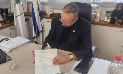 יצחק רוכברגר יושב ליד שולחן העבודה עם מסמך ביד אחת ועט ביד שנייה. על השולחן מסמכים, מקלדת, טלפון, סמארטפון, ובקבוק מים.