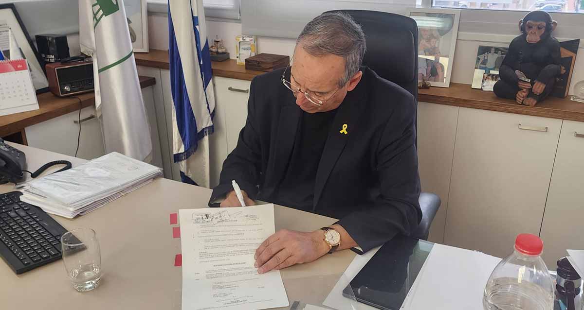 יצחק רוכברגר יושב ליד שולחן העבודה עם מסמך ביד אחת ועט ביד שנייה. על השולחן מסמכים, מקלדת, טלפון, סמארטפון, ובקבוק מים.