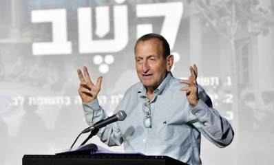 רון חולדאי לפני דוכן נואמים עם מיקרופון, ושתי ידיו מורמות והאצבעות כלפי מעלה. מאחוריו מוקרנת המילה: קשב