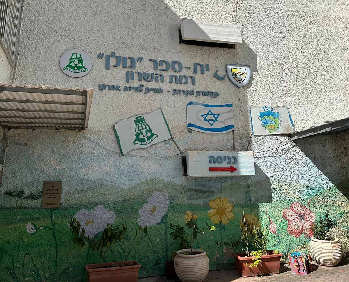 בית הספר היסודי גולן במערב רמת השרון. תמונה של קיר הכניסה לבית הספרץ למעלה הכיתוב: בי-ספר "גולן" רמת השרון, תקשורת מקרבת - חוויית למידה אחרת! על הקיר דגל המדינה, סמל העיר רמת השרון. חץ אדום המכוון לכניסה מימין. הקיר למטה עם ציור של פרחים וירק. לאורך הקיר מונחים עציצים עם צמחים.